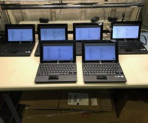 A row of seven laptops with their screens displaying system information, arranged on a workbench, likely for inventory or setup purposes.
