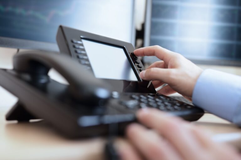 A hand operates a modern telephone on a desk, with digital screens displaying graphs in the background, indicating a business or financial context.
